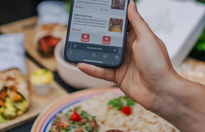 Hand&nbsp;holding&nbsp;a&nbsp;smartphone&nbsp;displaying&nbsp;a&nbsp;food&nbsp;delivery&nbsp;app&nbsp;screen&nbsp;in&nbsp;a&nbsp;restaurant&nbsp;setting.