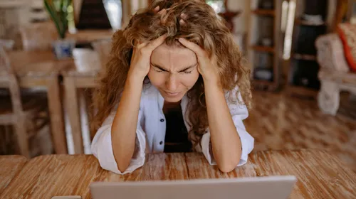 A&nbsp;stressed&nbsp;woman&nbsp;sits&nbsp;at&nbsp;a&nbsp;wooden&nbsp;table,&nbsp;frustrated&nbsp;with&nbsp;her&nbsp;laptop&nbsp;work.