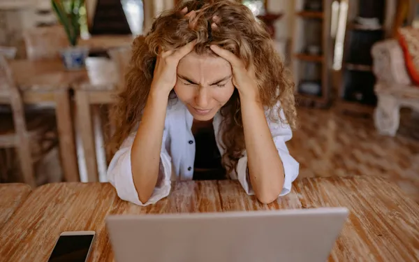 A&nbsp;stressed&nbsp;woman&nbsp;sits&nbsp;at&nbsp;a&nbsp;wooden&nbsp;table,&nbsp;frustrated&nbsp;with&nbsp;her&nbsp;laptop&nbsp;work.