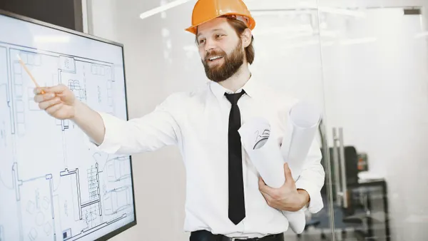 Engineer&nbsp;in&nbsp;helmet&nbsp;presenting&nbsp;architectural&nbsp;plans&nbsp;on&nbsp;a&nbsp;whiteboard&nbsp;in&nbsp;an&nbsp;office&nbsp;setting.