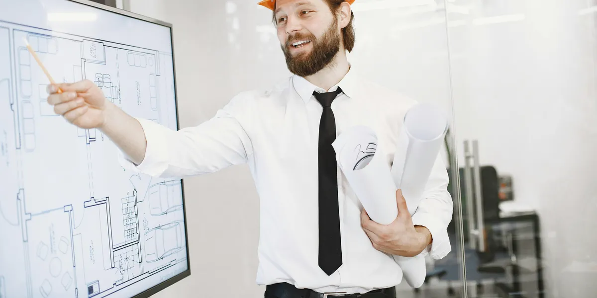 Engineer&nbsp;in&nbsp;helmet&nbsp;presenting&nbsp;architectural&nbsp;plans&nbsp;on&nbsp;a&nbsp;whiteboard&nbsp;in&nbsp;an&nbsp;office&nbsp;setting.