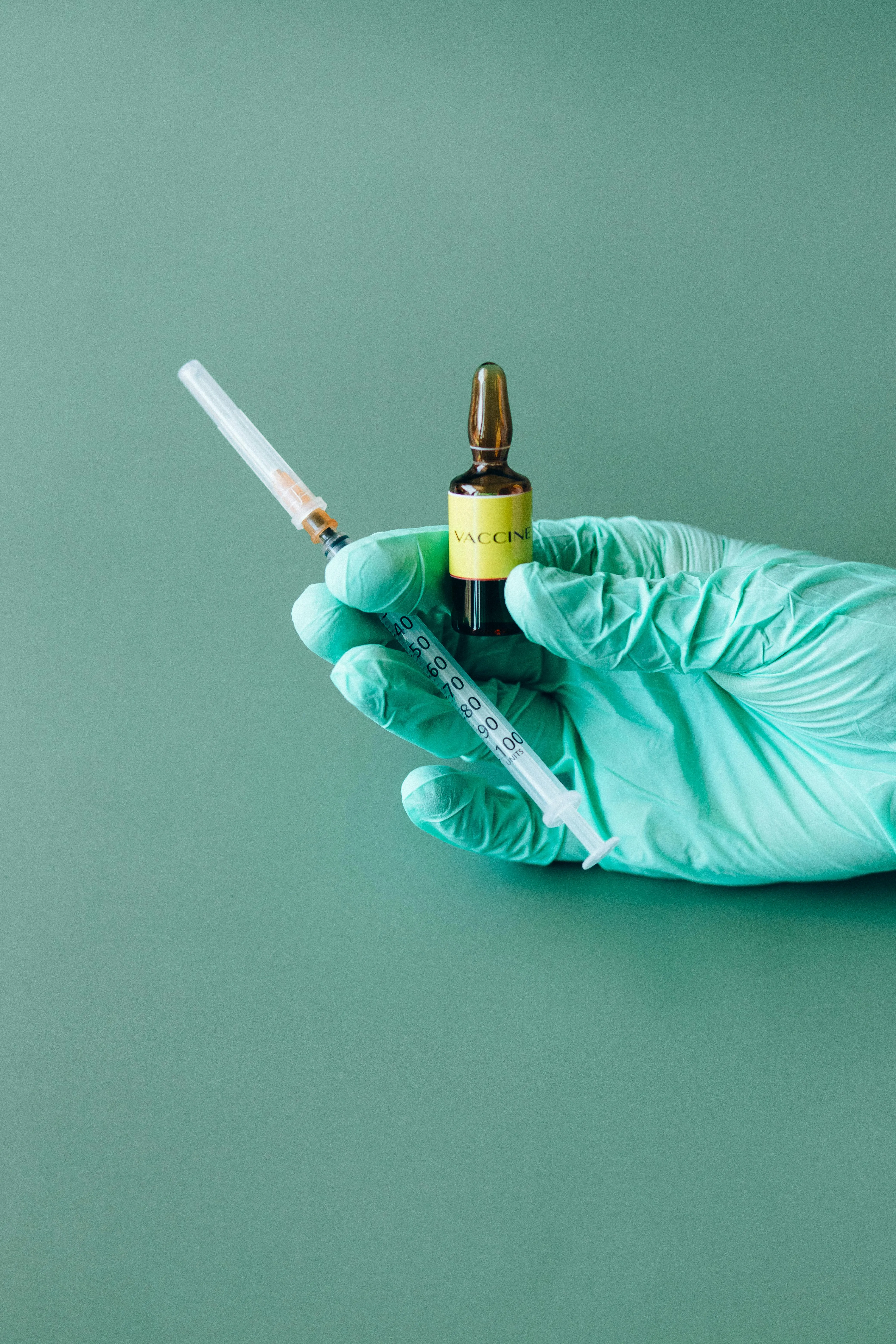 Close-up&nbsp;of&nbsp;a&nbsp;gloved&nbsp;hand&nbsp;holding&nbsp;a&nbsp;vaccine&nbsp;ampule&nbsp;and&nbsp;syringe&nbsp;on&nbsp;a&nbsp;green&nbsp;background.