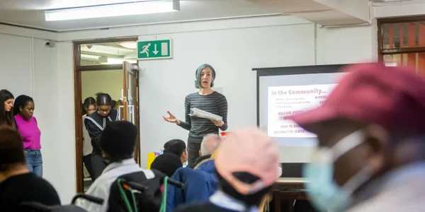 A&nbsp;diverse&nbsp;group&nbsp;attending&nbsp;a&nbsp;community&nbsp;meeting&nbsp;with&nbsp;a&nbsp;presenter&nbsp;speaking&nbsp;in&nbsp;front&nbsp;of&nbsp;a&nbsp;screen.