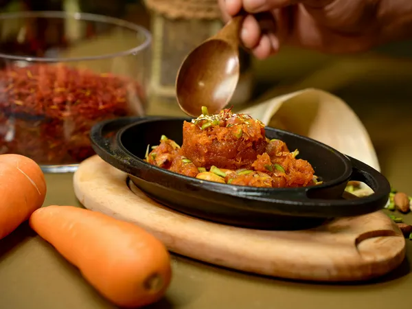 A&nbsp;vibrant&nbsp;Gajar&nbsp;Halwa&nbsp;garnished&nbsp;with&nbsp;pistachios,&nbsp;served&nbsp;in&nbsp;a&nbsp;black&nbsp;dish&nbsp;with&nbsp;carrots&nbsp;and&nbsp;saffron.