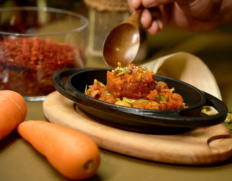 A&nbsp;vibrant&nbsp;Gajar&nbsp;Halwa&nbsp;garnished&nbsp;with&nbsp;pistachios,&nbsp;served&nbsp;in&nbsp;a&nbsp;black&nbsp;dish&nbsp;with&nbsp;carrots&nbsp;and&nbsp;saffron.