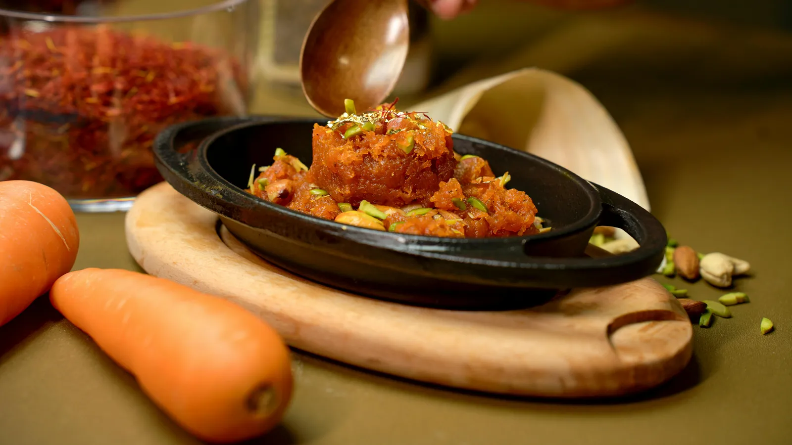 A&nbsp;vibrant&nbsp;Gajar&nbsp;Halwa&nbsp;garnished&nbsp;with&nbsp;pistachios,&nbsp;served&nbsp;in&nbsp;a&nbsp;black&nbsp;dish&nbsp;with&nbsp;carrots&nbsp;and&nbsp;saffron.