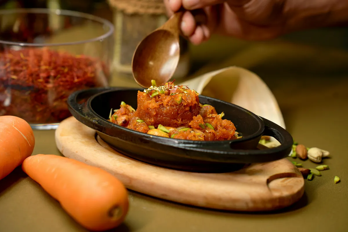 A&nbsp;vibrant&nbsp;Gajar&nbsp;Halwa&nbsp;garnished&nbsp;with&nbsp;pistachios,&nbsp;served&nbsp;in&nbsp;a&nbsp;black&nbsp;dish&nbsp;with&nbsp;carrots&nbsp;and&nbsp;saffron.