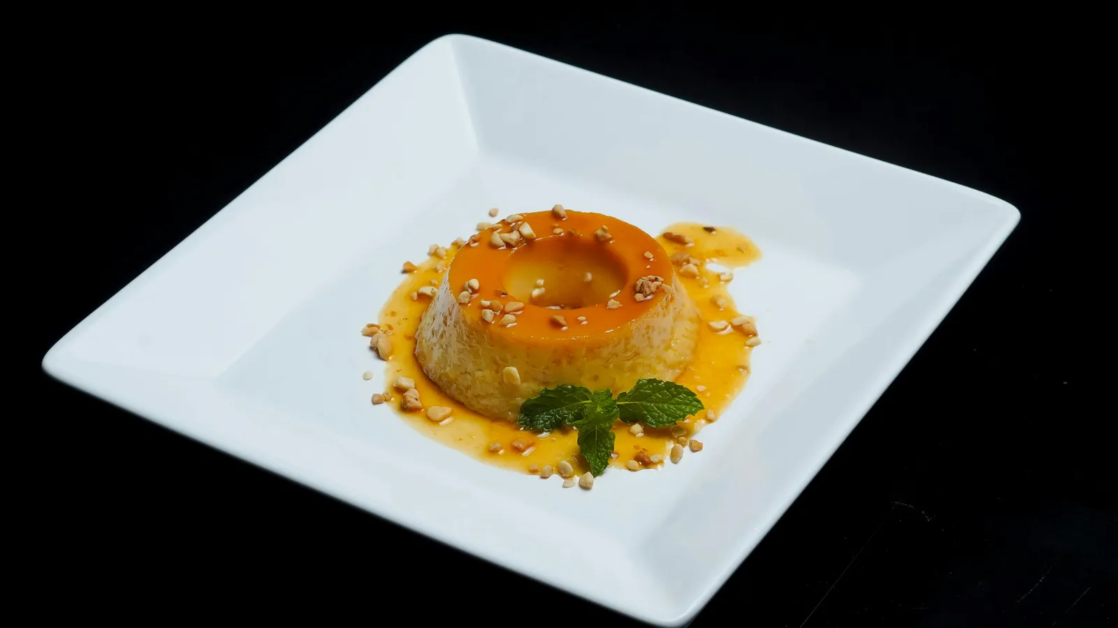 A&nbsp;beautifully&nbsp;plated&nbsp;caramel&nbsp;flan&nbsp;with&nbsp;mint&nbsp;leaves&nbsp;and&nbsp;nuts,&nbsp;set&nbsp;against&nbsp;a&nbsp;black&nbsp;background.